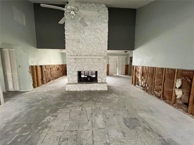 unfurnished living room with a towering ceiling, a wainscoted wall, ceiling fan, and a stone fireplace