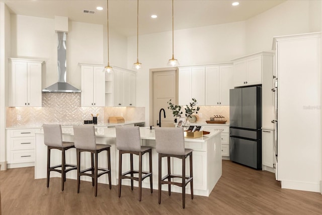 kitchen featuring a center island with sink, white cabinets, wall chimney exhaust hood, freestanding refrigerator, and open shelves