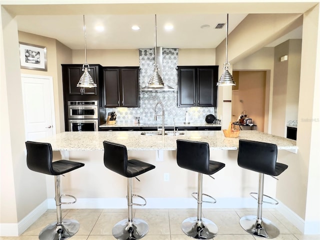 kitchen with wall chimney range hood, light stone countertops, a sink, a kitchen breakfast bar, and backsplash