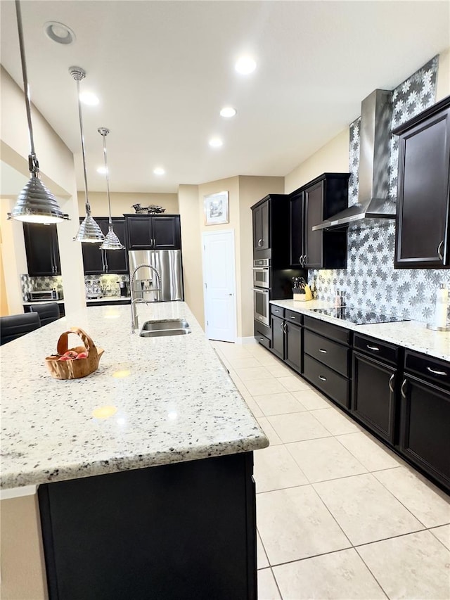 kitchen with a sink, light stone counters, appliances with stainless steel finishes, wall chimney exhaust hood, and dark cabinets