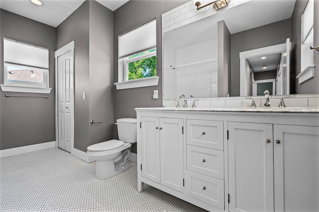 bathroom with baseboards, double vanity, a tile shower, a sink, and toilet