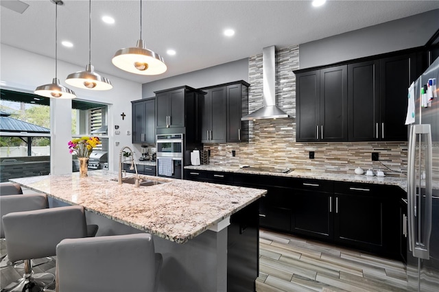 kitchen with tasteful backsplash, stainless steel appliances, a sink, wall chimney range hood, and dark cabinetry