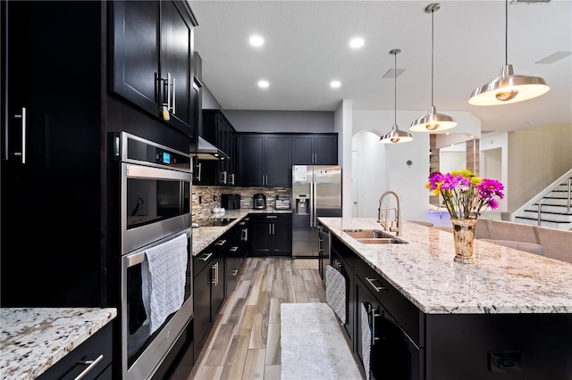 kitchen featuring a center island with sink, decorative backsplash, dark cabinets, stainless steel appliances, and a sink