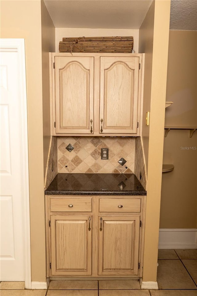 kitchen featuring dark stone countertops, light tile patterned floors, baseboards, light brown cabinetry, and backsplash