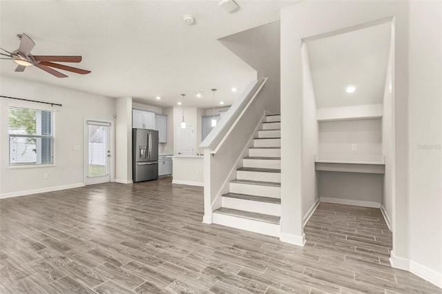 unfurnished living room with ceiling fan, light wood-style flooring, baseboards, and stairs