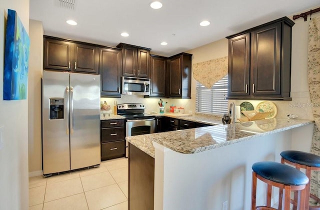 kitchen featuring light stone countertops, a peninsula, dark brown cabinets, appliances with stainless steel finishes, and a kitchen breakfast bar