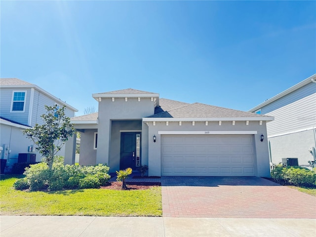 prairie-style house with stucco siding, central air condition unit, decorative driveway, a shingled roof, and a garage