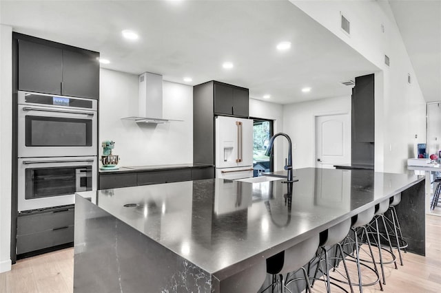 kitchen with stainless steel double oven, high end white refrigerator, light wood-style flooring, a sink, and wall chimney exhaust hood