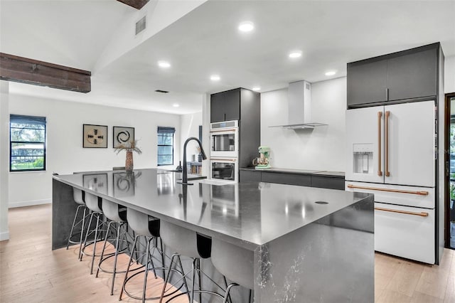 kitchen featuring white fridge with ice dispenser, wall chimney exhaust hood, light wood-style flooring, a spacious island, and double oven
