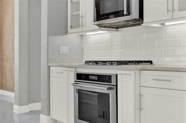 kitchen featuring light stone counters, stainless steel appliances, white cabinetry, backsplash, and glass insert cabinets