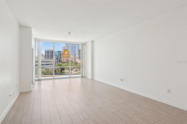 empty room with floor to ceiling windows, a city view, light wood-style flooring, and baseboards