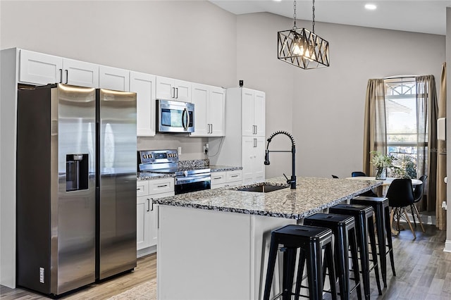 kitchen with light stone counters, a kitchen island with sink, a sink, stainless steel appliances, and white cabinetry