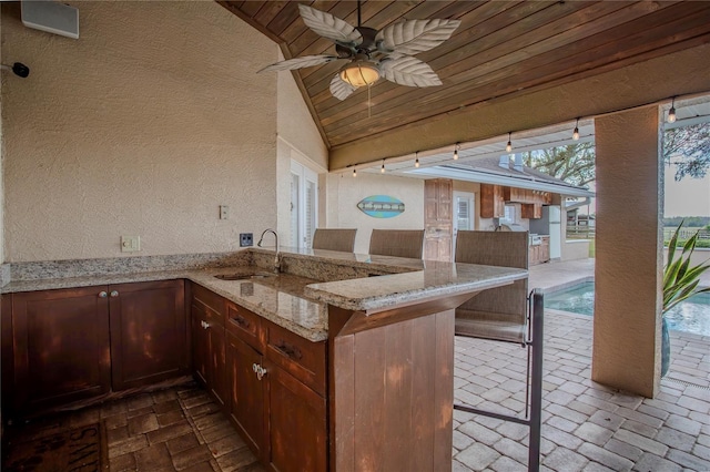 kitchen with a sink, light stone counters, a peninsula, brick floor, and wood ceiling