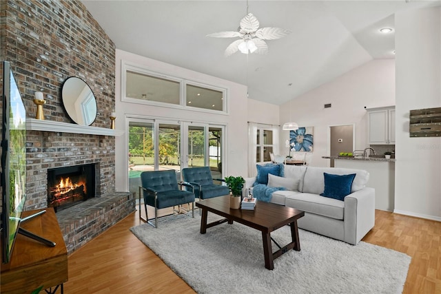 living room with a brick fireplace, high vaulted ceiling, light wood finished floors, and ceiling fan