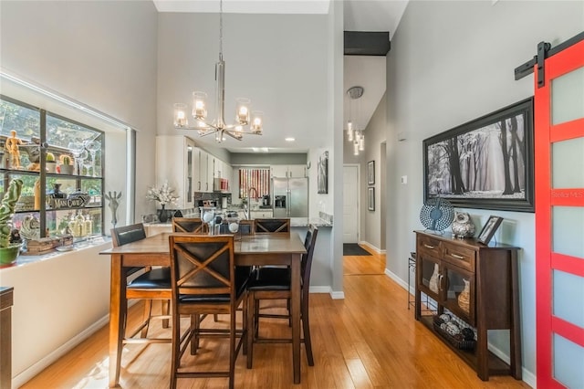 dining area with a notable chandelier, light wood-style flooring, a towering ceiling, a barn door, and baseboards