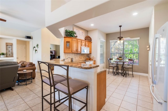 kitchen with light stone countertops, a breakfast bar area, light tile patterned floors, white appliances, and a ceiling fan