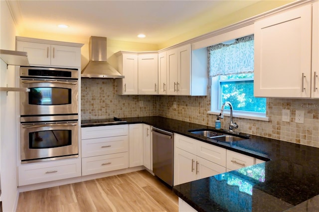 kitchen featuring a sink, light wood-style floors, appliances with stainless steel finishes, white cabinets, and wall chimney range hood