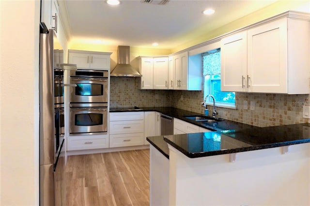 kitchen with light wood-style flooring, a sink, a peninsula, appliances with stainless steel finishes, and wall chimney exhaust hood