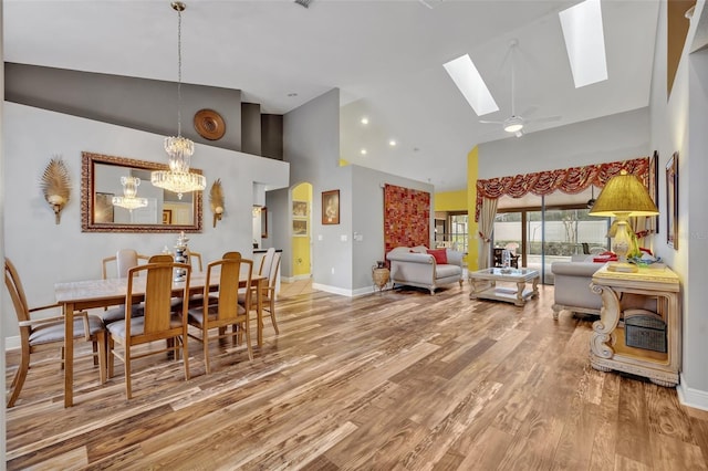 dining area with a skylight, baseboards, light wood-type flooring, high vaulted ceiling, and ceiling fan with notable chandelier