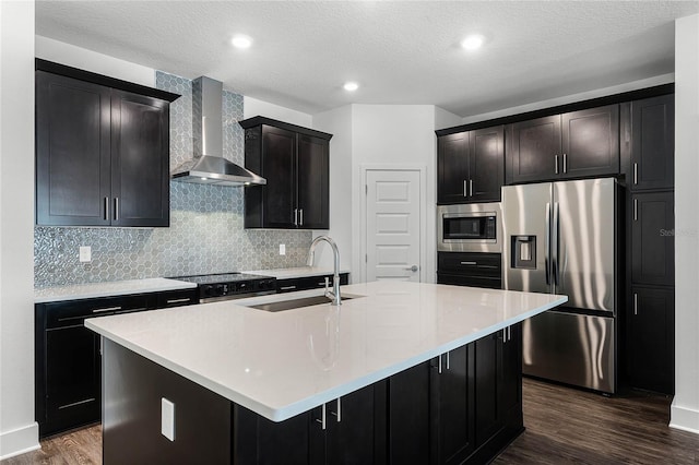 kitchen featuring dark wood-style floors, appliances with stainless steel finishes, a sink, an island with sink, and wall chimney exhaust hood