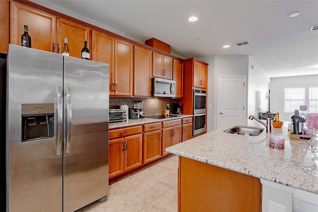 kitchen with light stone counters, backsplash, appliances with stainless steel finishes, brown cabinetry, and a sink