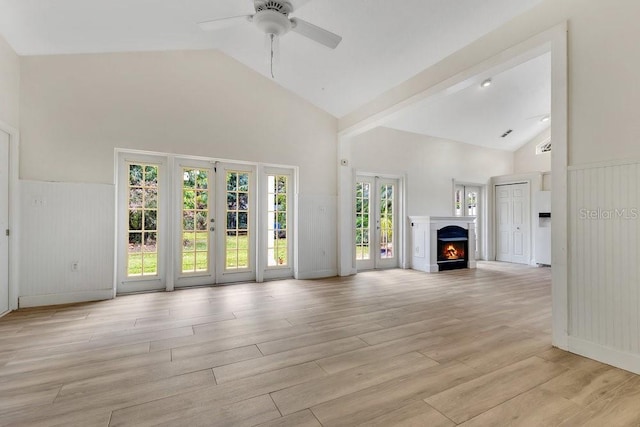 unfurnished living room featuring wainscoting, light wood-style flooring, french doors, a warm lit fireplace, and high vaulted ceiling