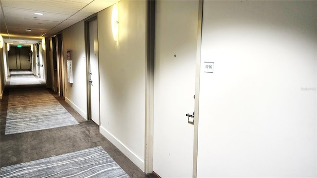 corridor with a paneled ceiling, baseboards, and dark carpet