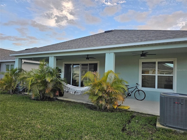 back of house with stucco siding, a ceiling fan, central AC, a yard, and a shingled roof