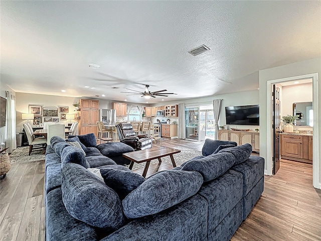 living area featuring light wood-type flooring, ceiling fan, visible vents, and a textured ceiling