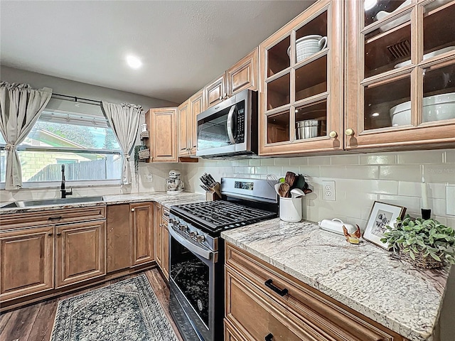 kitchen with light stone counters, a sink, appliances with stainless steel finishes, backsplash, and glass insert cabinets