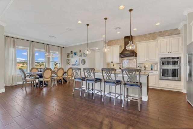 kitchen featuring tasteful backsplash, a kitchen bar, custom exhaust hood, stainless steel double oven, and dark wood-style flooring