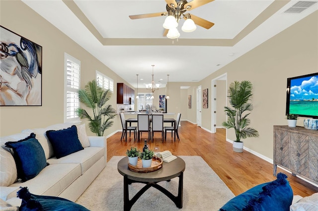 living area with visible vents, light wood-style flooring, ceiling fan with notable chandelier, a tray ceiling, and baseboards