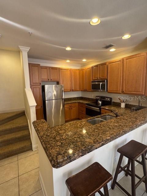 kitchen with stainless steel appliances, a peninsula, a sink, brown cabinets, and dark stone countertops