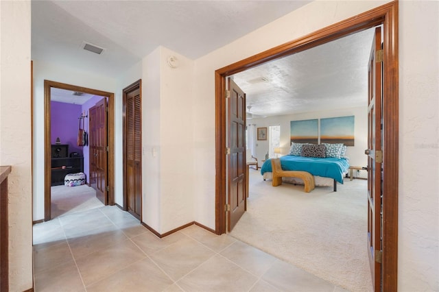 hallway featuring visible vents, light carpet, light tile patterned flooring, baseboards, and a textured wall