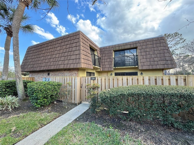 view of side of property with brick siding, mansard roof, fence, and a balcony