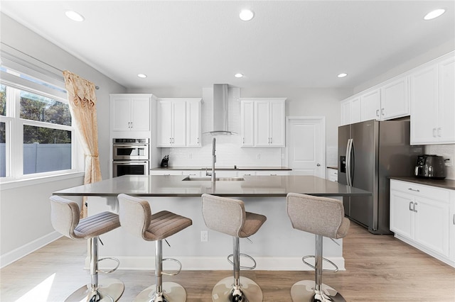 kitchen featuring dark countertops, stainless steel appliances, light wood-style floors, wall chimney exhaust hood, and decorative backsplash