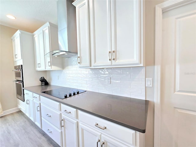 kitchen featuring stainless steel double oven, white cabinets, wall chimney exhaust hood, black electric cooktop, and backsplash