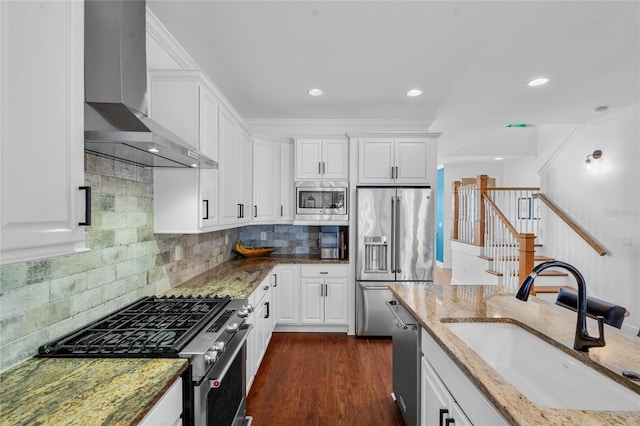 kitchen featuring light stone counters, appliances with stainless steel finishes, white cabinets, wall chimney exhaust hood, and a sink