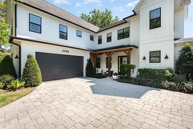 modern inspired farmhouse with a shingled roof, stucco siding, a garage, decorative driveway, and board and batten siding