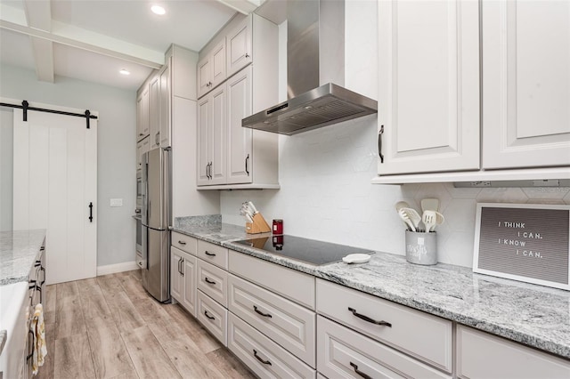 kitchen featuring backsplash, a barn door, light wood-style floors, wall chimney exhaust hood, and black electric stovetop