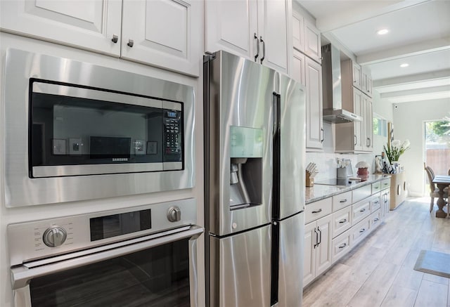 kitchen featuring light wood finished floors, white cabinets, appliances with stainless steel finishes, wall chimney range hood, and backsplash