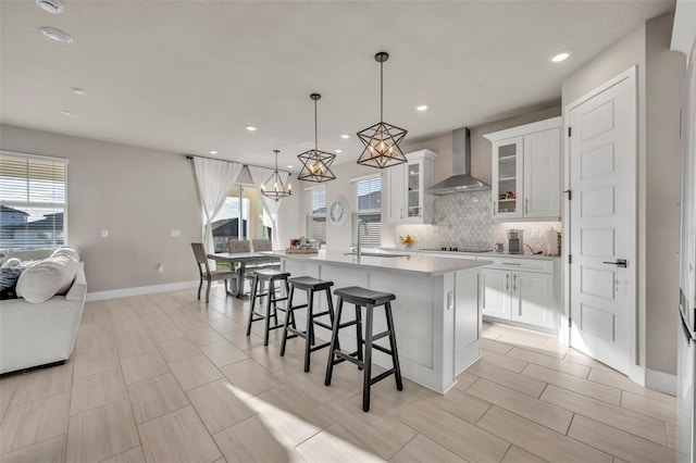 kitchen with a kitchen island with sink, decorative backsplash, light countertops, white cabinetry, and wall chimney exhaust hood