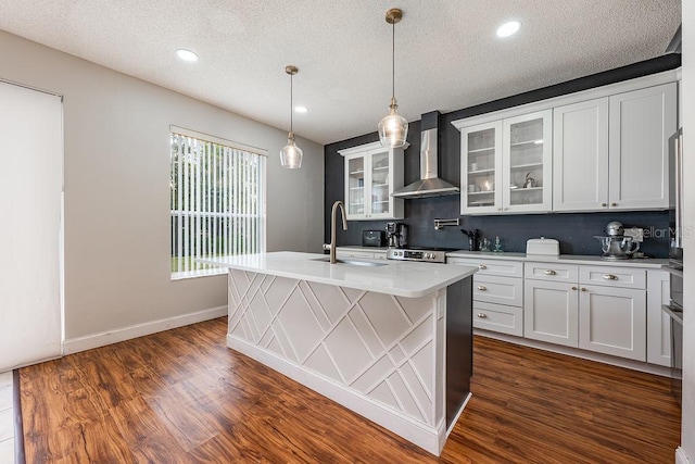 kitchen with an island with sink, light countertops, dark wood-style floors, wall chimney exhaust hood, and a sink