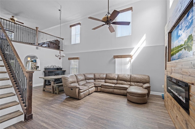 living room with wood finished floors, baseboards, stairs, a glass covered fireplace, and ceiling fan with notable chandelier