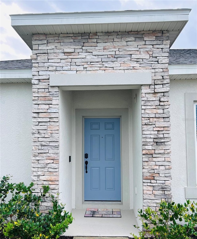 property entrance with stucco siding, stone siding, and roof with shingles