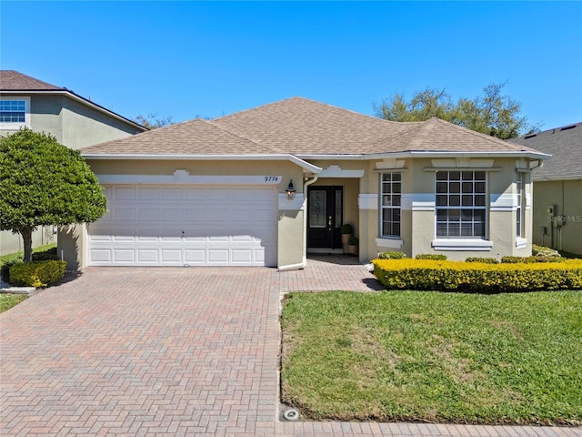 ranch-style house with a front yard, an attached garage, a shingled roof, stucco siding, and decorative driveway