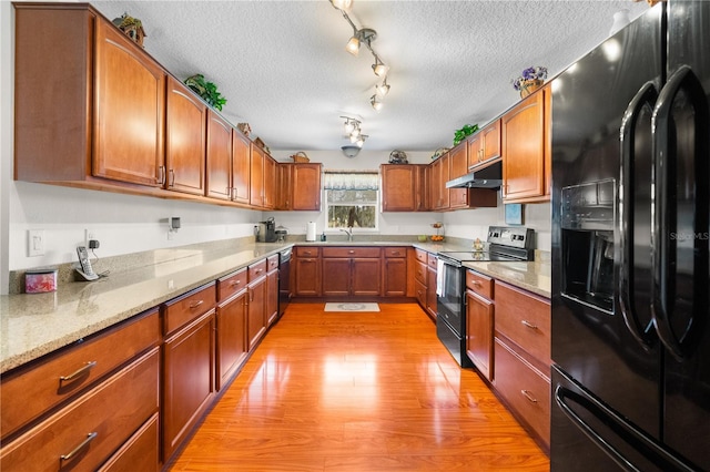 kitchen featuring light wood finished floors, under cabinet range hood, brown cabinetry, black appliances, and a sink