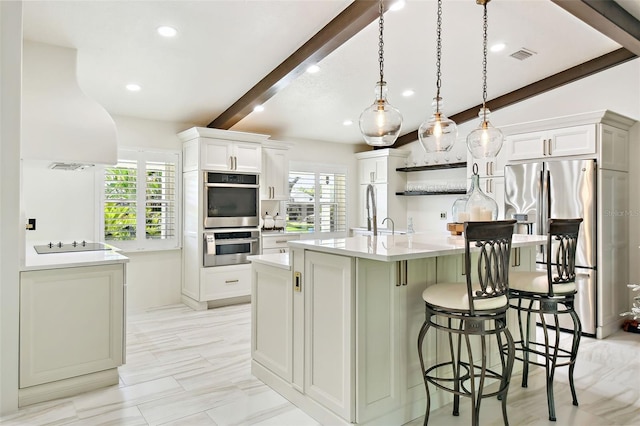 kitchen featuring beamed ceiling, a center island with sink, a kitchen bar, light countertops, and appliances with stainless steel finishes