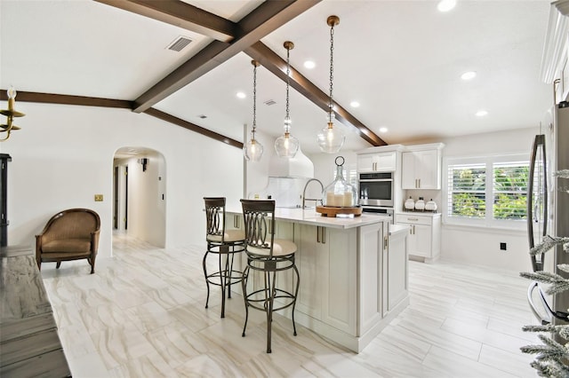 kitchen with vaulted ceiling with beams, a center island with sink, double oven, a kitchen bar, and arched walkways