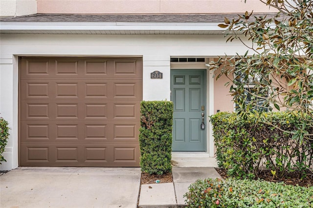 entrance to property featuring concrete driveway, a garage, roof with shingles, and stucco siding